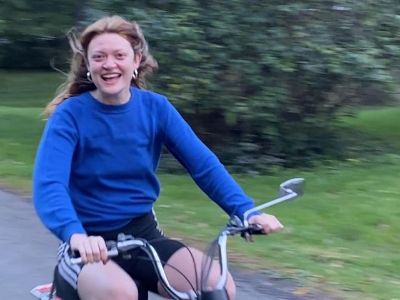 Colby Minifie is wearing a blue top as she smiles at the camera while riding a moped.
